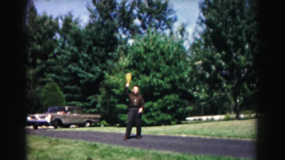 A man in dark clothing handles a yellow flying disc in a 1963 park scene, with a period car, lawn, path, and trees in the background.