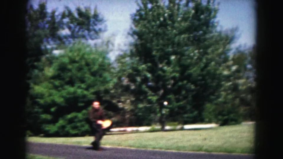 The player moves across the paved path with the yellow disc nearby in 1963 Hagerstown park footage, framed by lawns, trees, and 8mm vignette edges.