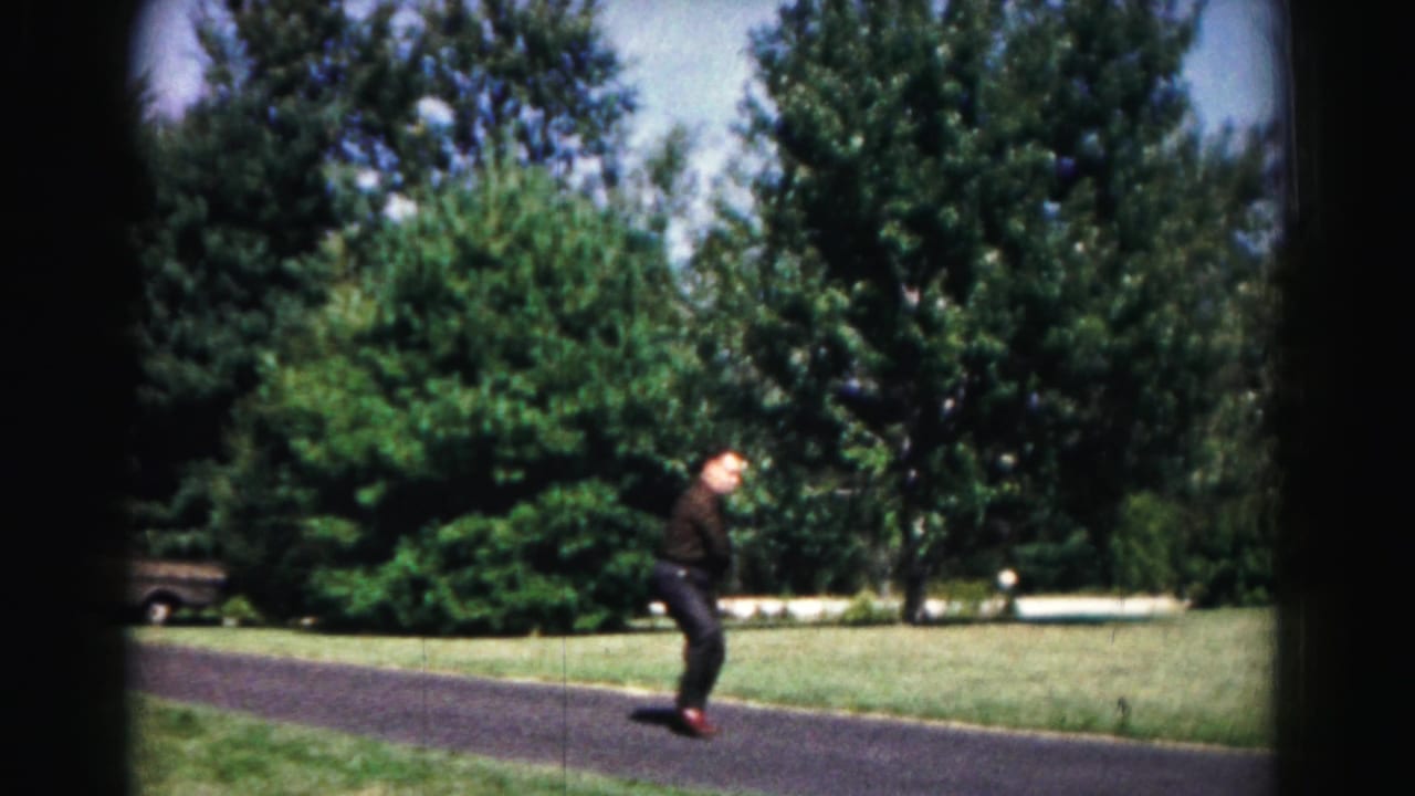 In 1963 Hagerstown home-movie footage, a young man in dark clothing waits with a racket on a paved path near lawn and trees.
