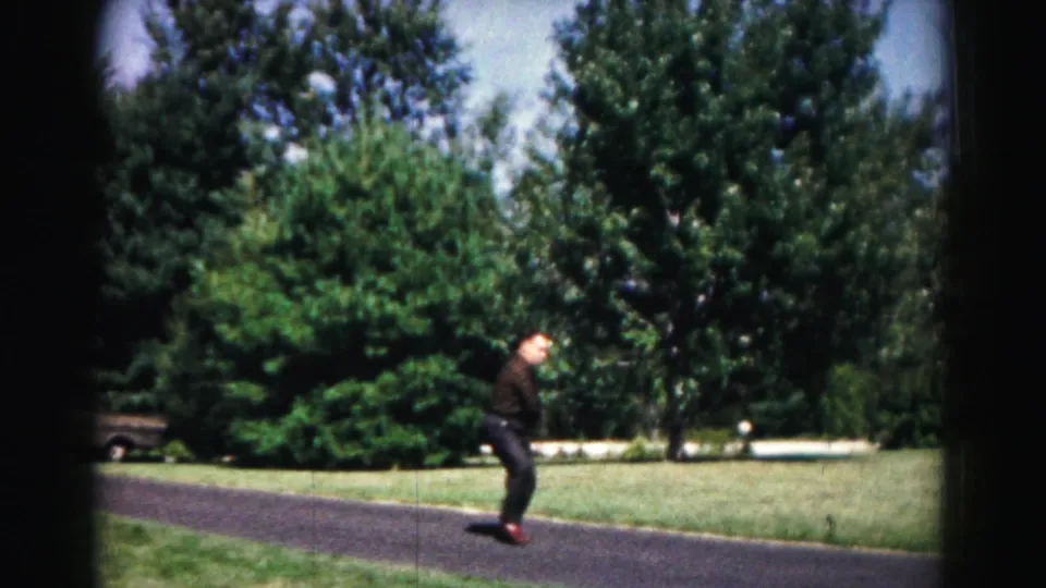 In 1963 Hagerstown home-movie footage, a young man in dark clothing waits with a racket on a paved path near lawn and trees.
