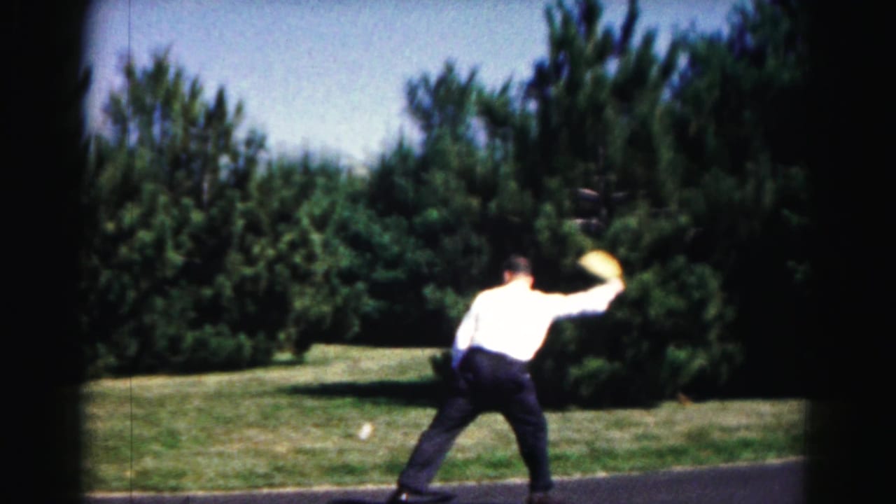 A young man in a white shirt swings a racket during street tennis in a sunny Hagerstown, Maryland outdoor setting.