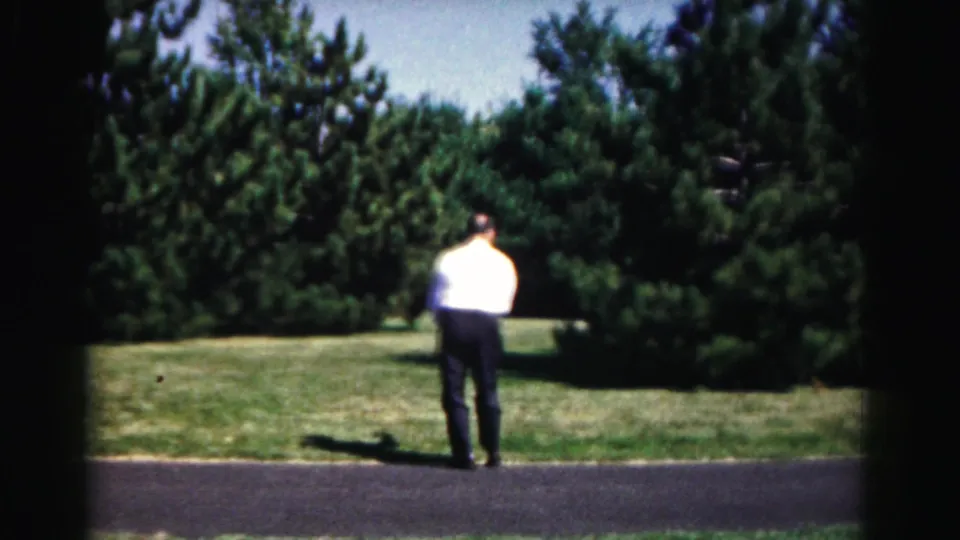 The player in a white shirt stands facing the trees and lawn at the end of the short 1963 Hagerstown street tennis moment.