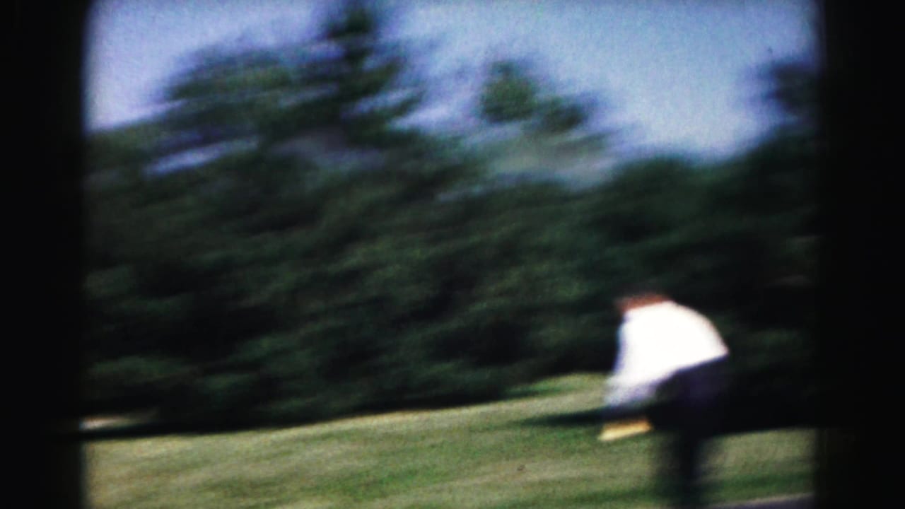 Blurred man in a white shirt practicing tennis on a grassy yard near trees.