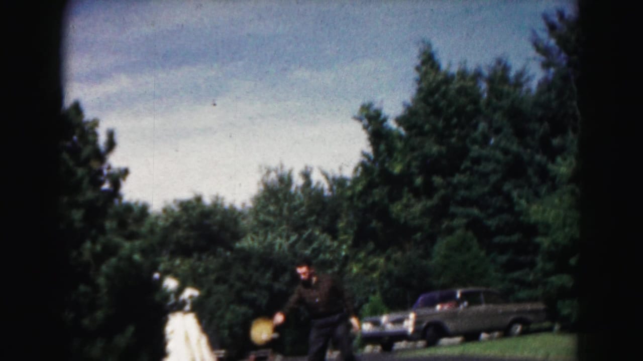 1963 Hagerstown home-movie frame showing two adults playing a casual yard game near trees and a parked sedan.