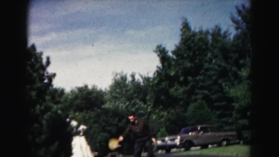 1963 Hagerstown home-movie frame showing two adults playing a casual yard game near trees and a parked sedan.