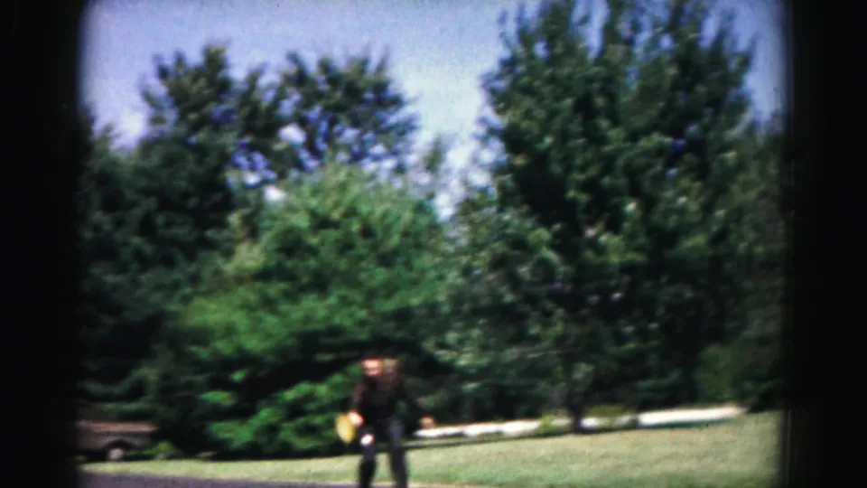 A player in dark clothing holds a paddle-like bat on a leafy 1963 Hagerstown lawn during the home-movie game.