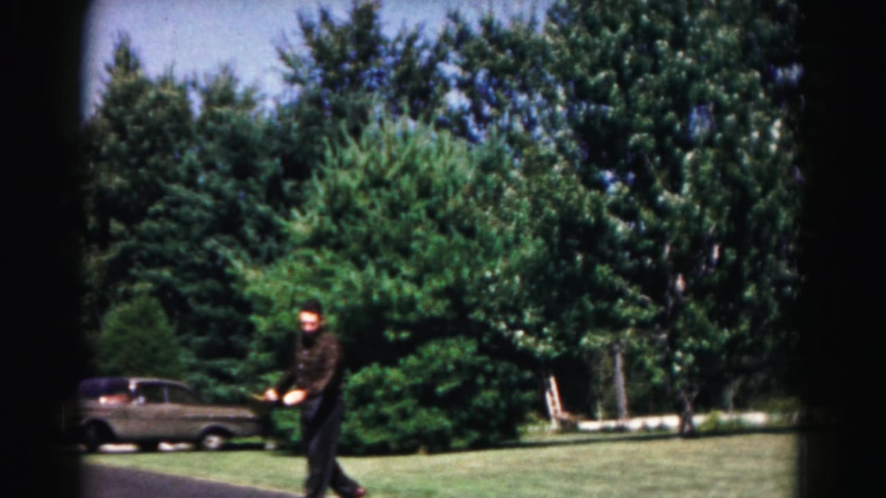 Man in dark clothing swings a paddle in a suburban yard with a parked car and trees behind him.