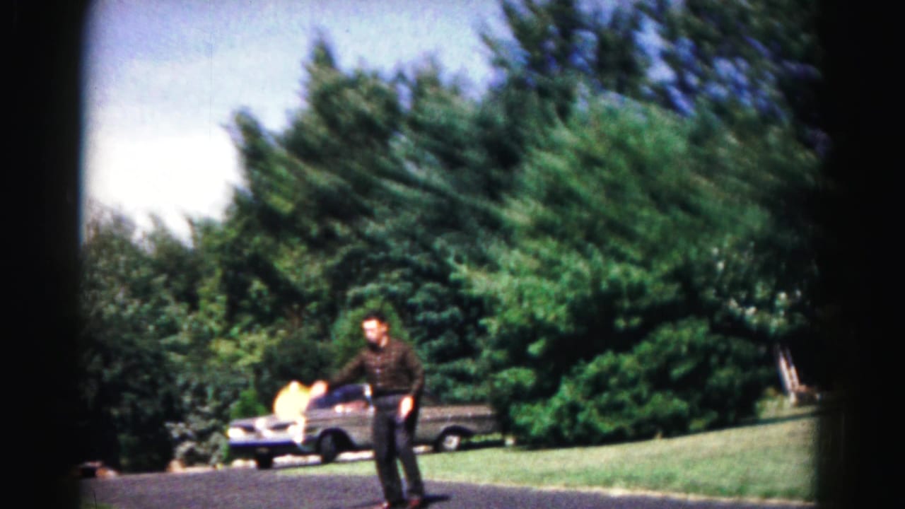 Man faces the camera while holding a paddle, with a ball blur, car, lawn, and trees in the background.