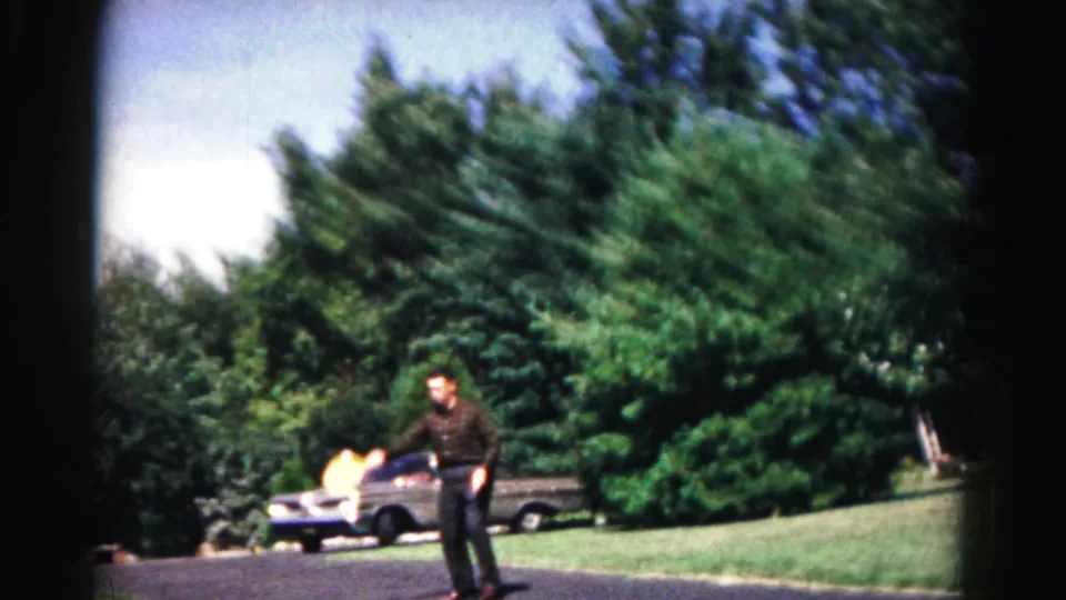 A man playing paddle ball stands in a Hagerstown suburban yard, with a light ball blur near the parked car and trees.