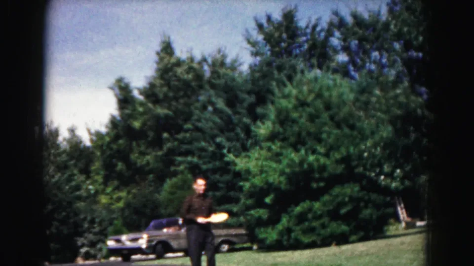 The 8mm frame shows the paddle-ball player in a suburban Maryland yard, with a mid-century car and heavy greenery behind him.