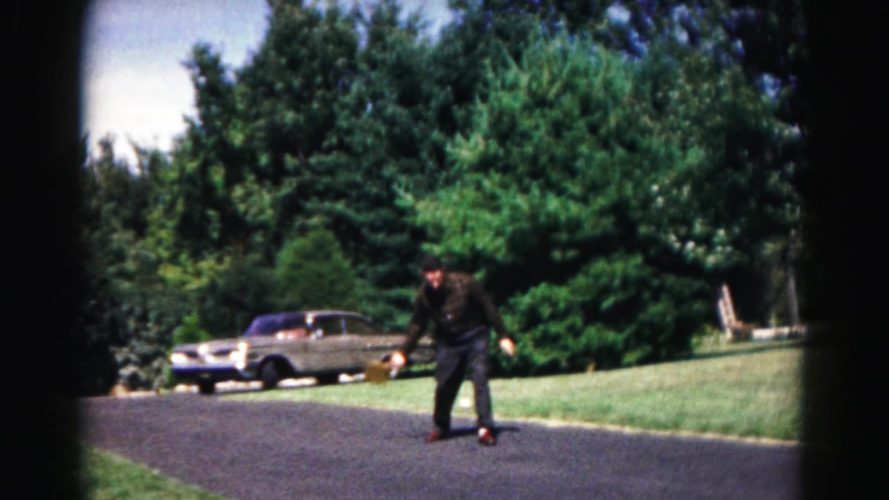 Man bends with a badminton racket on a sunny road near a parked sedan and trees.