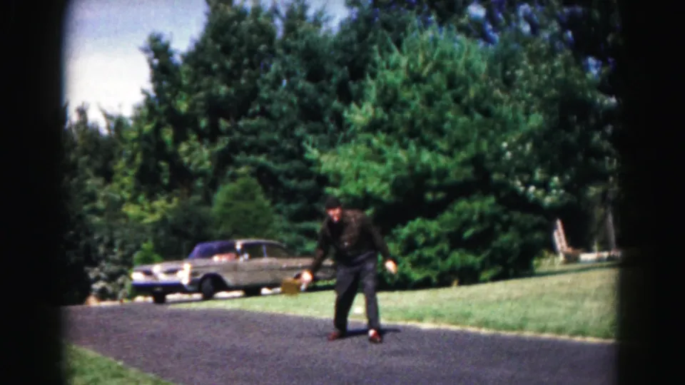 In 1963 Hagerstown, Maryland footage, a man with a racket bends on an asphalt road beside grass, trees, and a parked sedan.