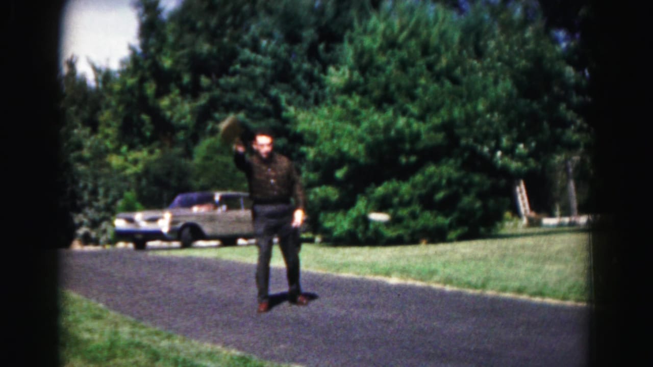 Man raises a badminton racket while standing on a road with a sedan parked behind him.