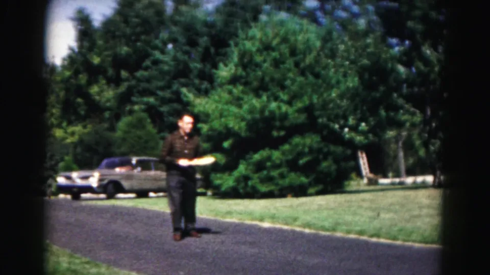 The man walks along the asphalt holding a badminton racket, framed by green lawn, trees, and a parked sedan in Hagerstown home-movie footage.