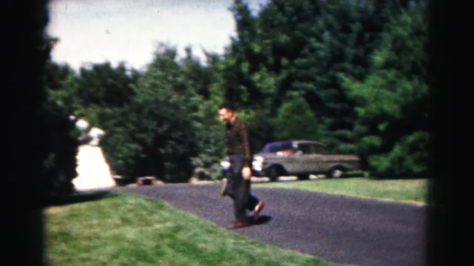 At the end of the clip, the man crosses the road with his racket while a parked sedan, grass, and dense trees remain in view.