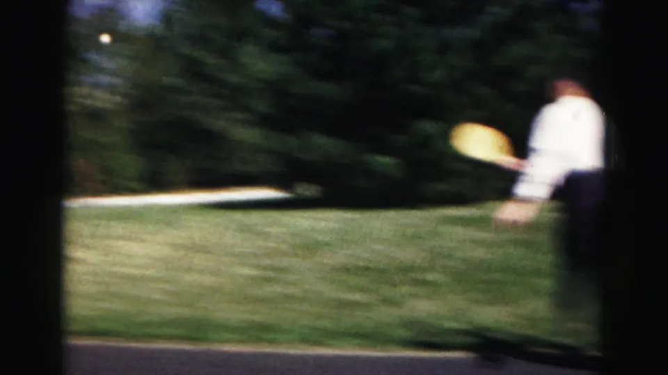 Soft, motion-blurred 1963 8mm frame from Hagerstown showing a person with a yellow disc beside a suburban lawn and driveway.