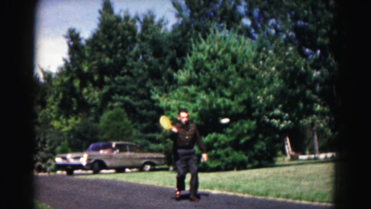 A man stands on a suburban driveway in Hagerstown, Maryland, holding a yellow disc near a parked mid-century sedan and dense trees.