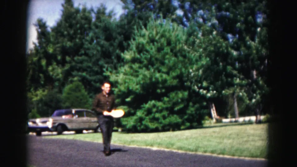 The man carries the yellow disc along the driveway, with a parked sedan, lawn, and trees visible in the 1963 suburban home-movie frame.