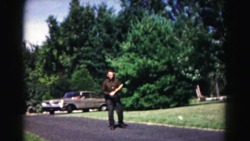 A man pauses on the driveway with the yellow disc in hand, framed by a lawn, dense trees, and a parked sedan in Hagerstown home-movie footage.