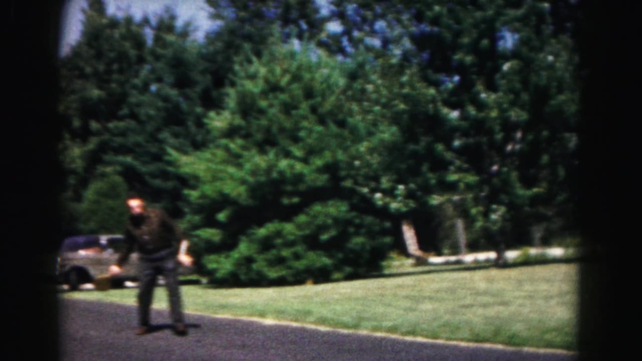 A man in dark clothing stands on a suburban driveway with a paddle, with lawn, trees, and a parked car behind him.