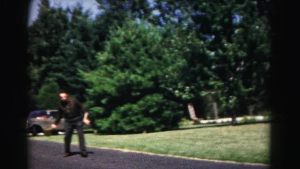A man in dark clothing stands on a suburban driveway with a paddle, with lawn, trees, and a parked car behind him.