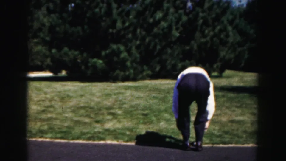A man in a white shirt bends at the edge of a suburban driveway, likely retrieving the ball during the 1963 paddleball game.