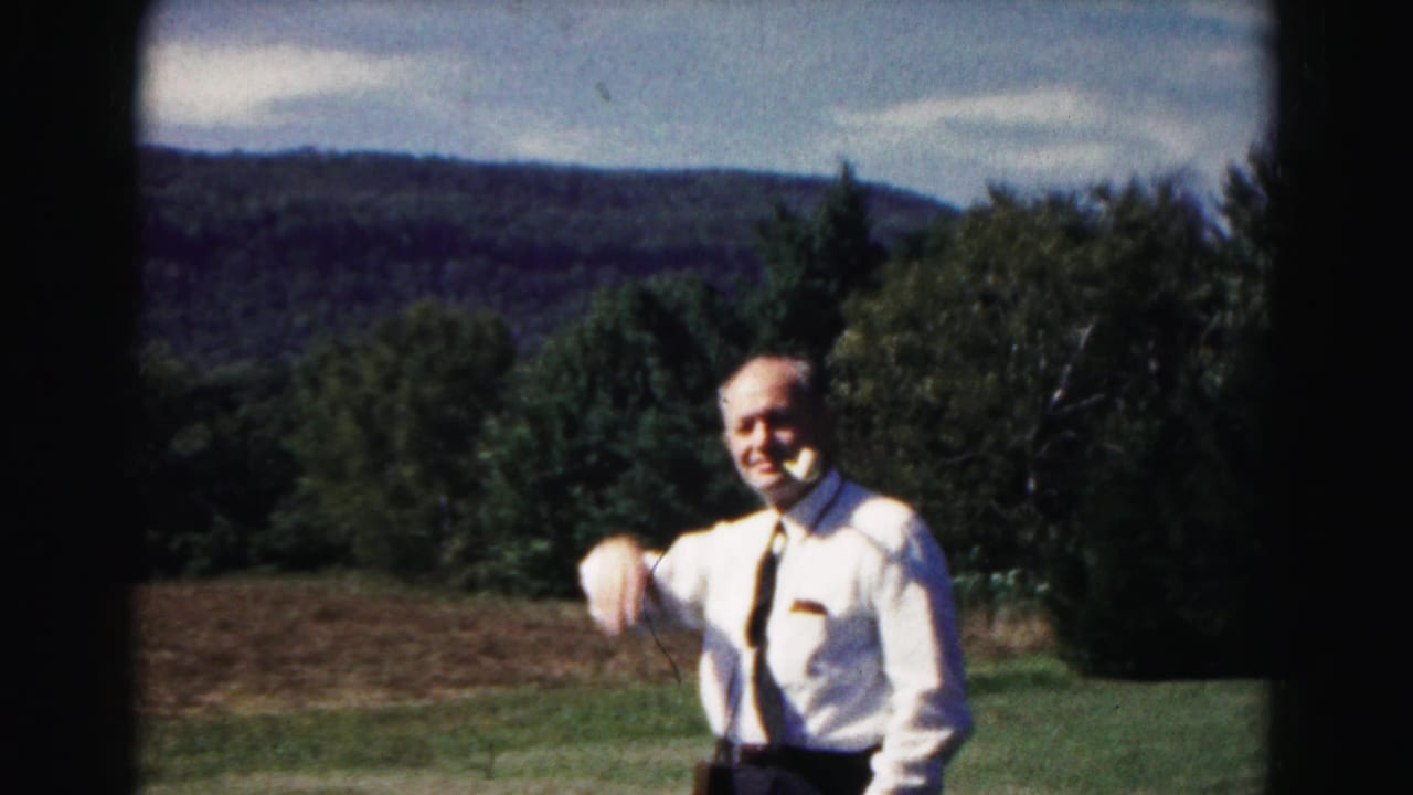 Smiling man in a white shirt and tie gesturing in a field with trees and hills behind him.