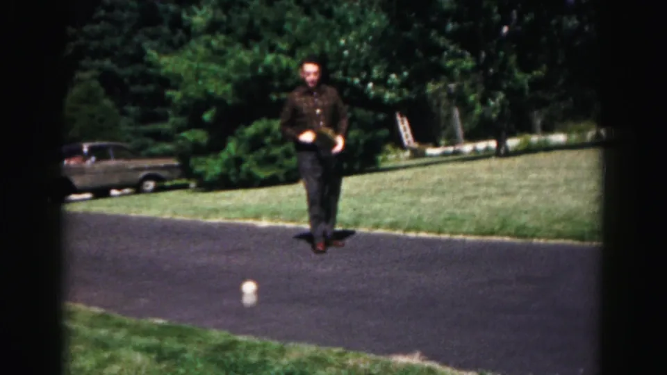 Adult man in a plaid shirt walks along a paved driveway in the 1963 Hagerstown-area home movie, with a parked older car nearby.