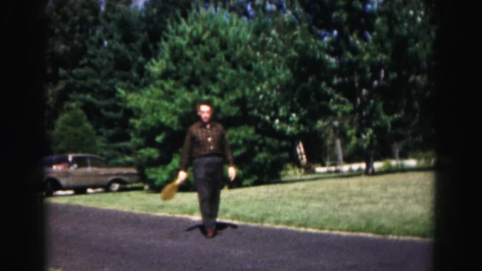 Adult man carries a yellow object while walking on a driveway in a green country setting, with trees and an older car in view.