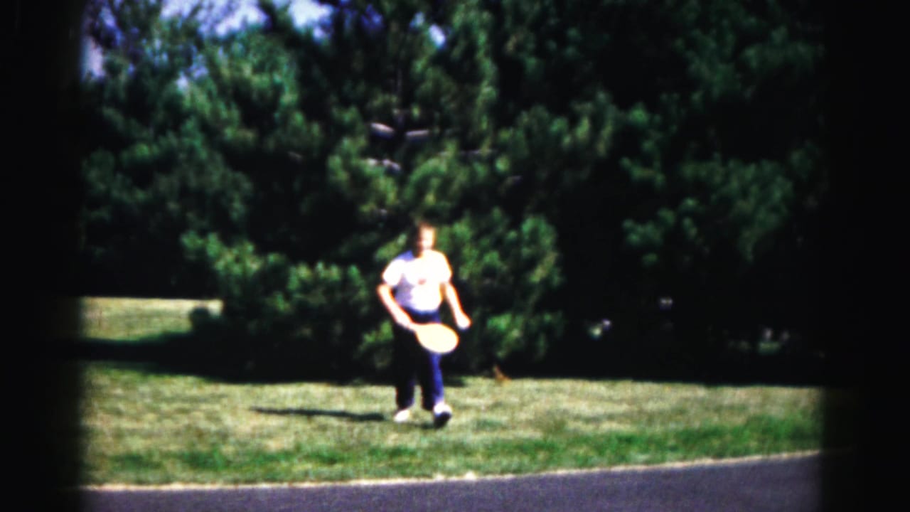1963 Hagerstown-area home-movie frame showing a man in a light shirt walking across grass near dense trees and a road edge.