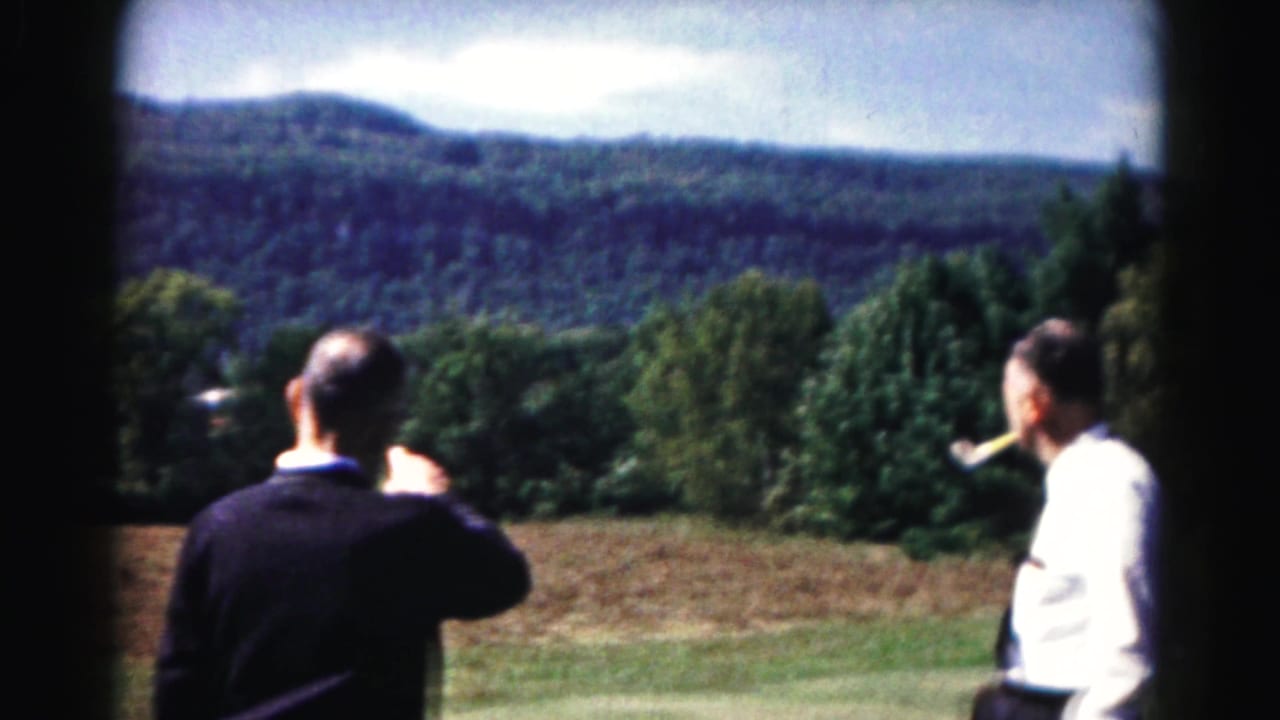 Two men stand in a field looking toward a wooded mountain landscape.