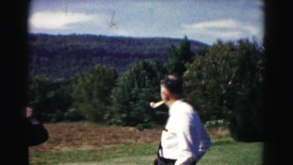 Home-movie frame of an adult man in a white shirt smoking a pipe near an open field, trees, and forested mountains in Maryland.