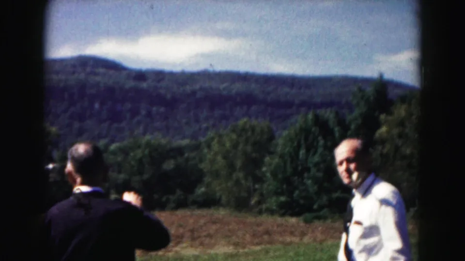 1963 home-movie frame near Hagerstown showing two men at a scenic overlook, one facing the camera while mountains and trees sit behind them.