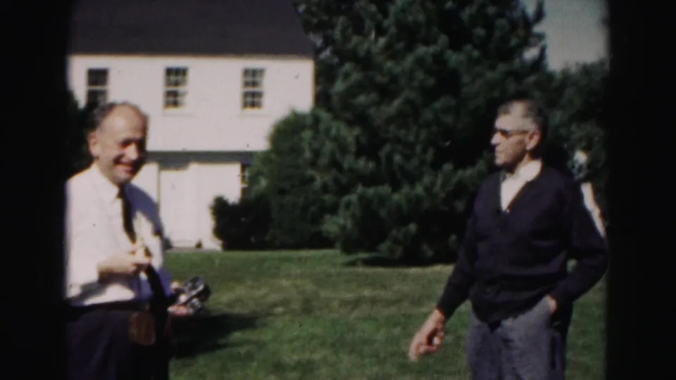 Two adult men appear on a residential lawn in 1963 Hagerstown, one smiling in a shirt and tie and the other wearing glasses and a dark cardigan.