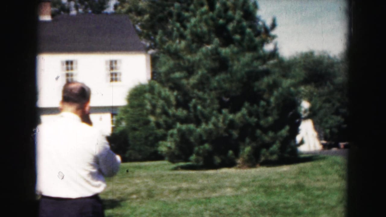In 1963 Hagerstown home-movie footage, a man in a white shirt stands on a lawn facing a white house framed by evergreens.