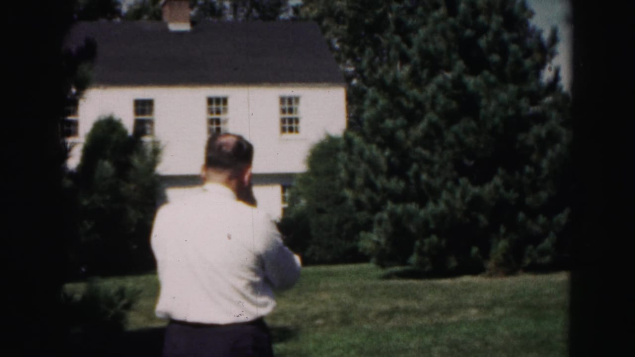 The man remains turned away from the camera in a 1963 Hagerstown garden scene, with a white house and dense evergreens beyond the lawn.