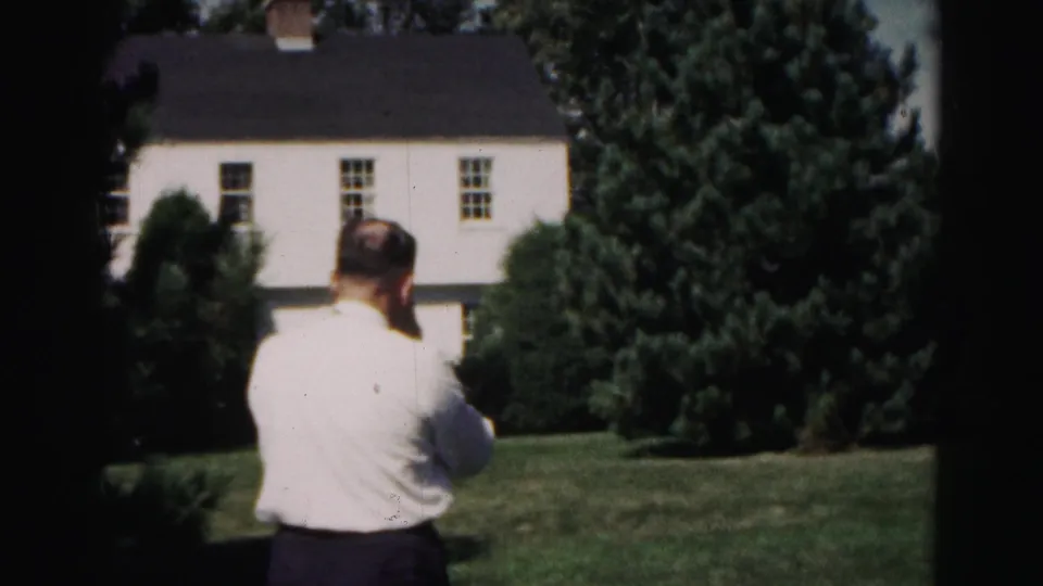 The man remains turned away from the camera in a 1963 Hagerstown garden scene, with a white house and dense evergreens beyond the lawn.