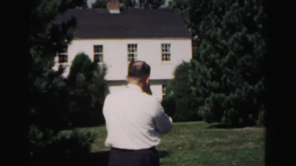 A 1963 Hagerstown home-movie frame shows the man from behind on the lawn, with evergreen trees and the white house filling the background.