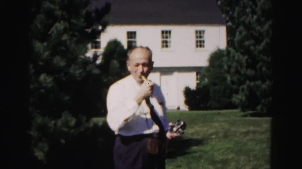 In the 1963 Hagerstown garden scene, the man turns toward the camera, smiling while holding a pipe, with the white house behind him.