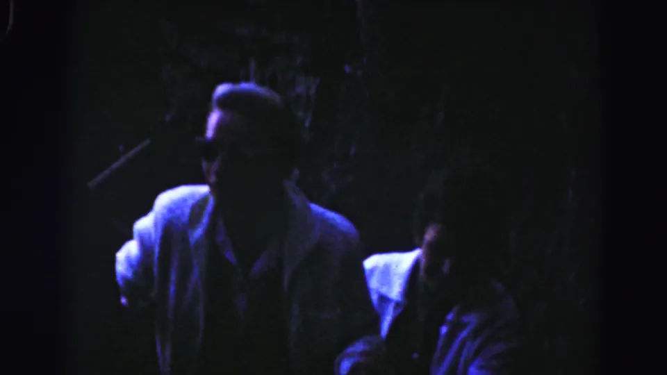 Two adult men pass close to the camera while ascending a dim stairway, with faces softened by shadow and blue-purple 8mm color.