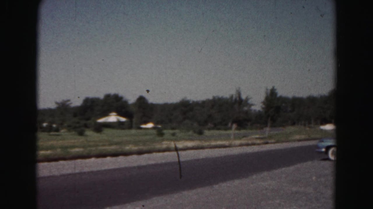 1963 8mm view near Hagerstown, Maryland, showing a rural road, grass, trees, and distant white shelters or tents.