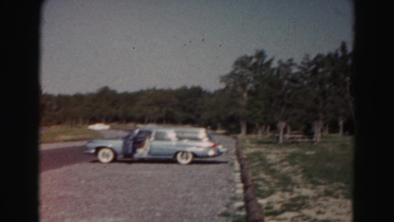 Light blue early-1960s station wagon parked in a gravel lot near grass, trees, and distant white shelters in Maryland home-movie footage.