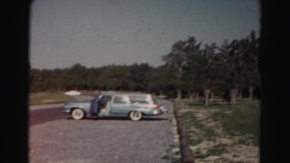 Light blue early-1960s station wagon parked in a gravel lot near grass, trees, and distant white shelters in Maryland home-movie footage.
