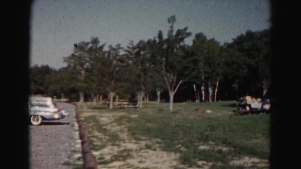Partial 1963 8mm frame of a parked station wagon at the edge of a gravel lot, with grass, trees, and a picnic table nearby.