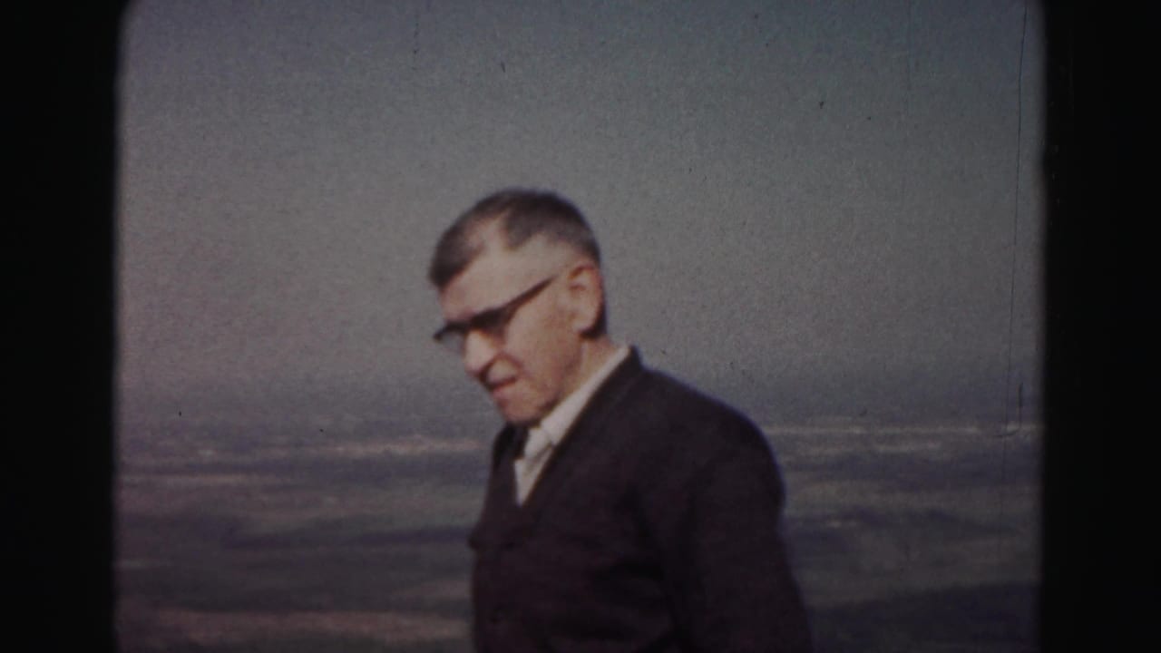 Adult man in glasses and dark jacket stands near a scenic overlook with distant landscape behind him.