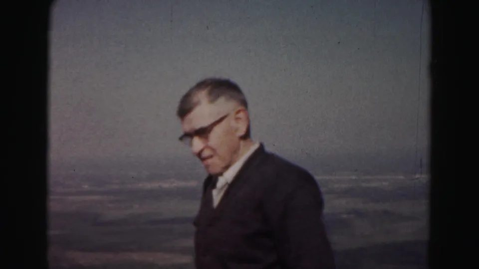 In 1963 Hagerstown, Maryland home-movie footage, an adult man in glasses stands at a high overlook with hazy fields in the distance.