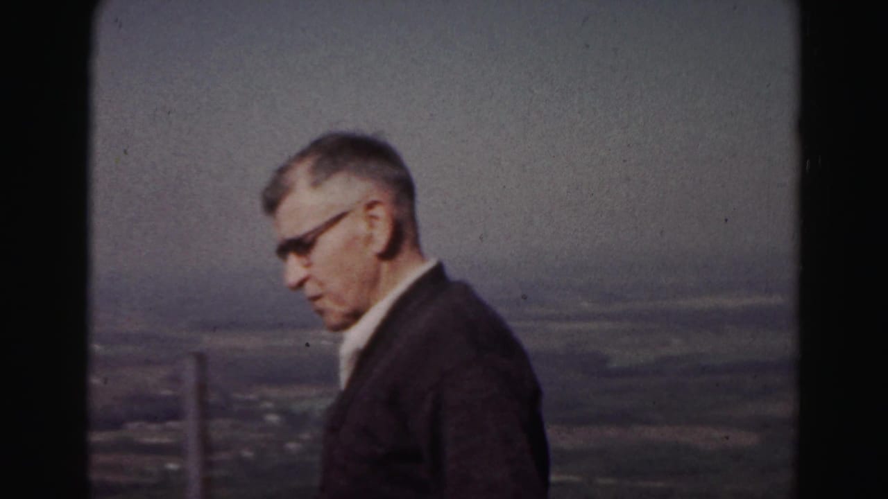 Adult man in profile stands by an overlook with a railing post and broad landscape behind him.