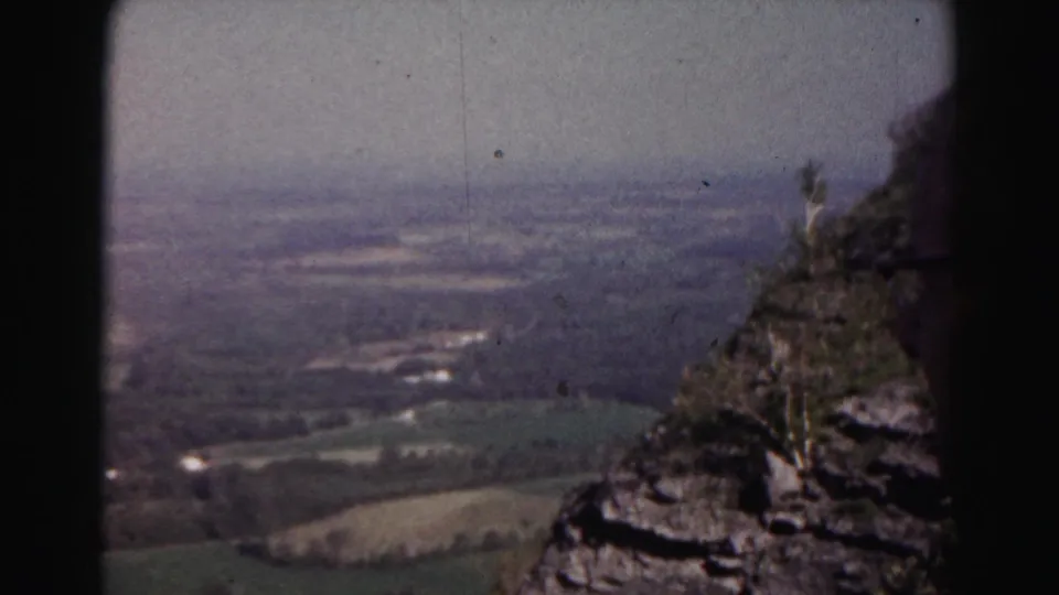 A 1963 8mm home-movie frame shows green fields and distant settlement below a rocky mountainside near Hagerstown, Maryland.