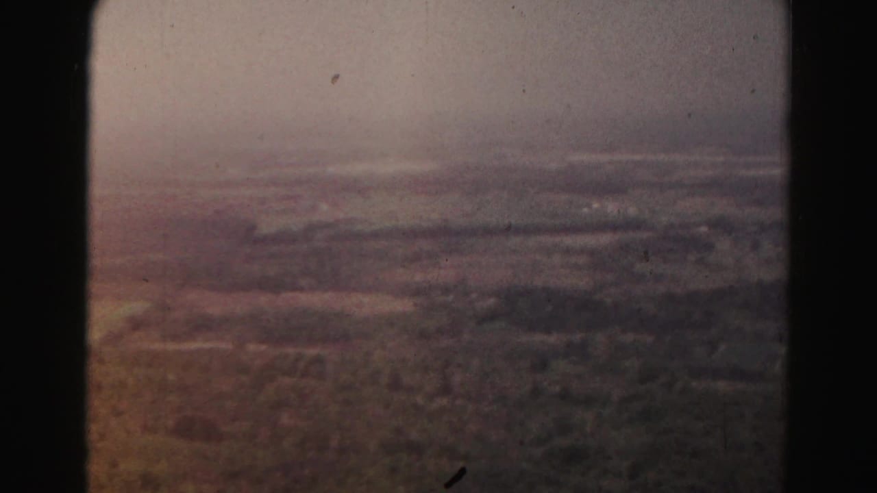 Soft aerial view of rural fields and distant haze, framed by dark film borders.