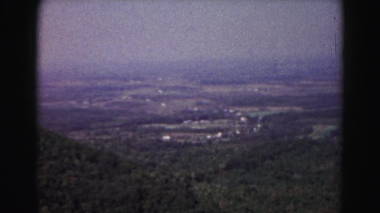 Forested valley and distant fields under a hazy sky, framed by dark 8mm film borders.